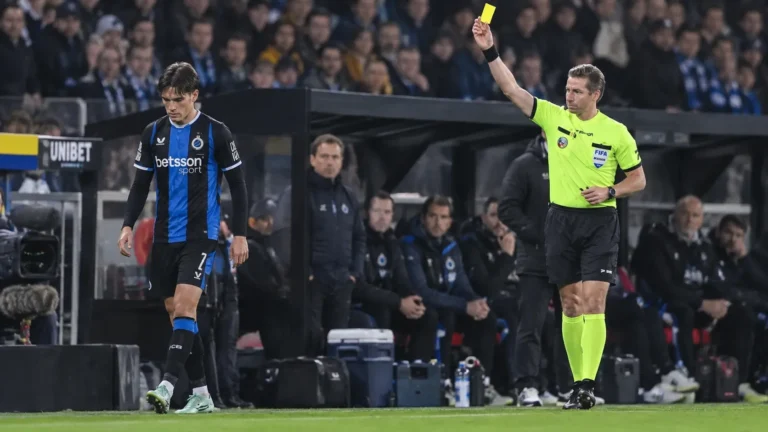 Een scheidsrechter toont een gele kaart aan een speler van Club Brugge naast de dug-out.