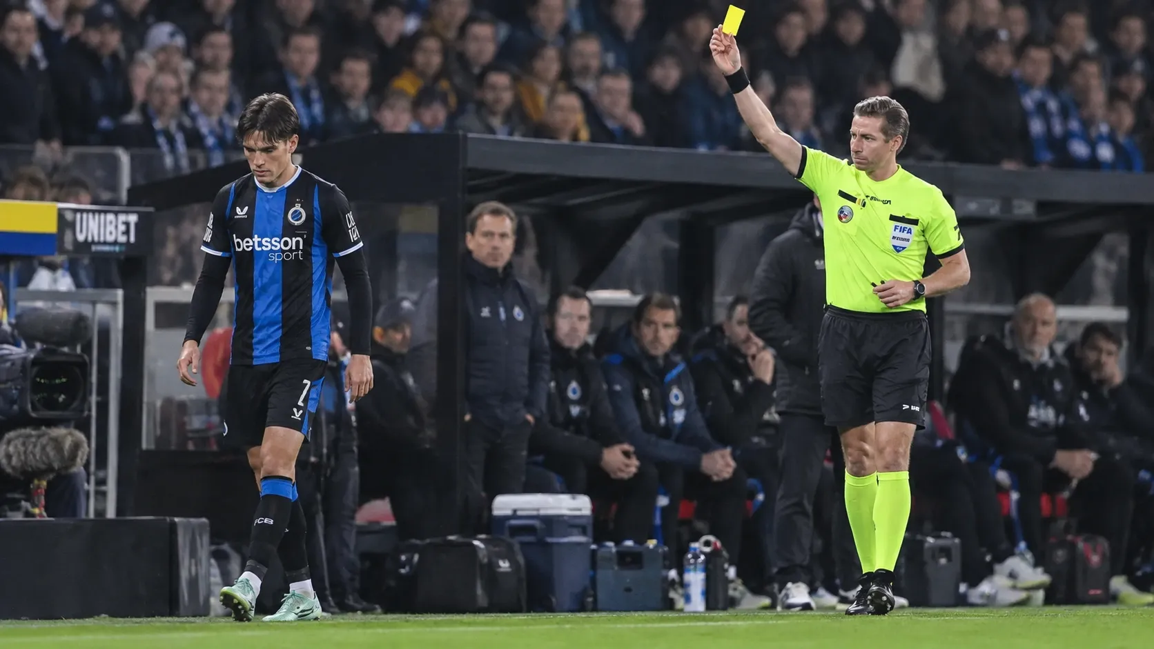 Een scheidsrechter toont een gele kaart aan een speler van Club Brugge naast de dug-out.