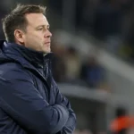 Een coach in een donkerblauwe jas aan de zijlijn van een voetbalstadion.
