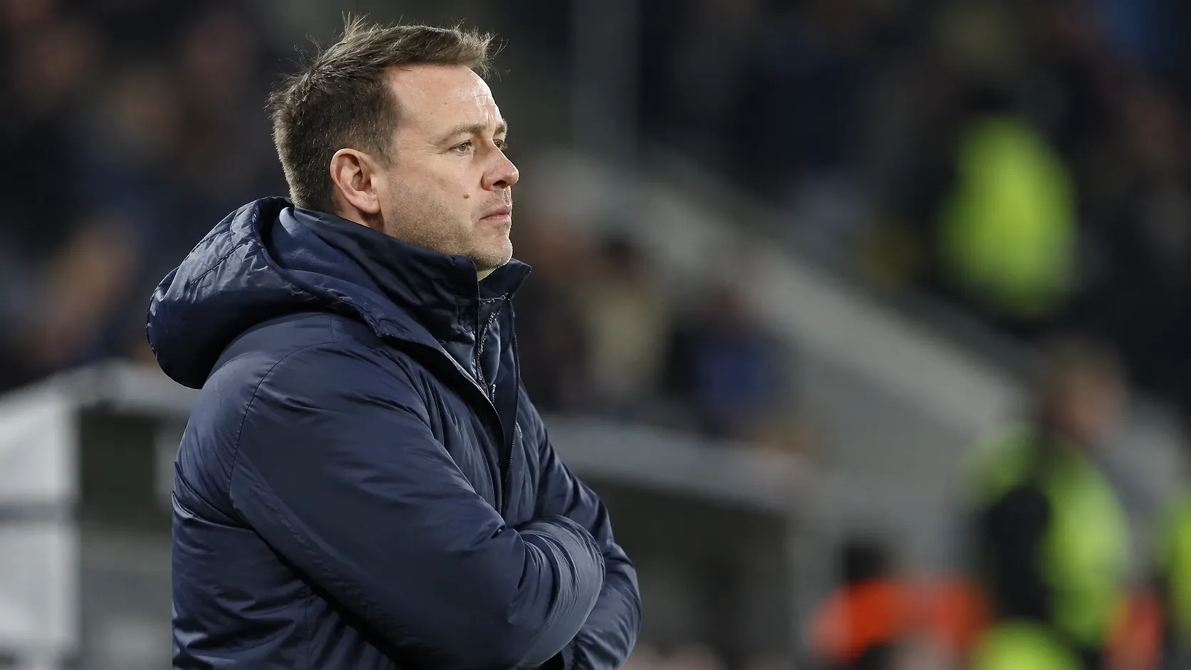 Een coach in een donkerblauwe jas aan de zijlijn van een voetbalstadion.