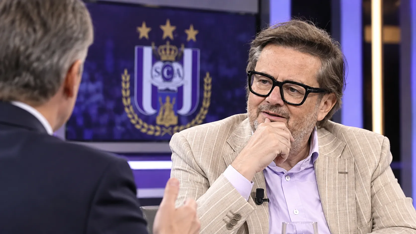 Marc Coucke spreekt in een televisiestudio met een subtiel RSC Anderlecht-embleem op een studiemonitor.