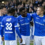 Een groep KRC Genk-spelers in blauwe wedstrijdkleding wandelt samen voorbij de tribune.