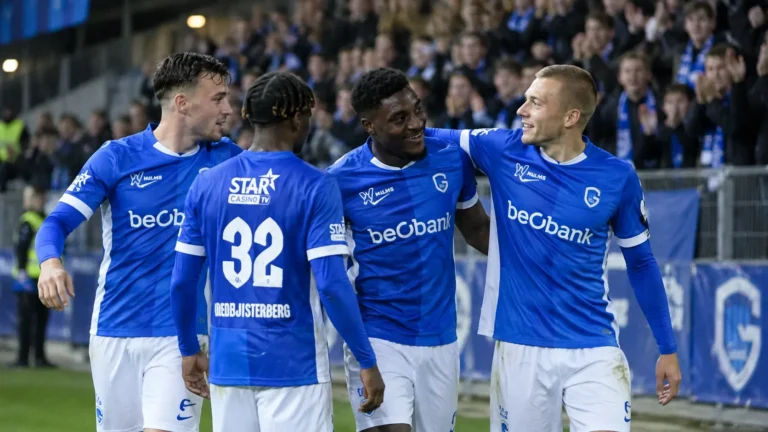 Een groep KRC Genk-spelers in blauwe wedstrijdkleding wandelt samen voorbij de tribune.