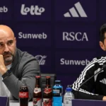 Twee deelnemers aan een persconferentie zitten achter een tafel met microfoons.