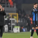 Een trainer in een zwarte jas en een speler in blauw-zwarte kleding applaudisseren langs het veld.
