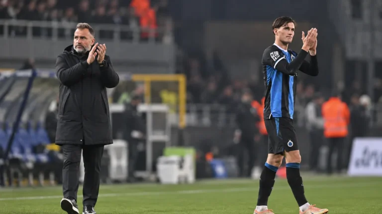 Een trainer in een zwarte jas en een speler in blauw-zwarte kleding applaudisseren langs het veld.