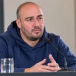 Stijn Stijnen spreekt in een blauwe hoodie aan een tafel tijdens een persconferentie.