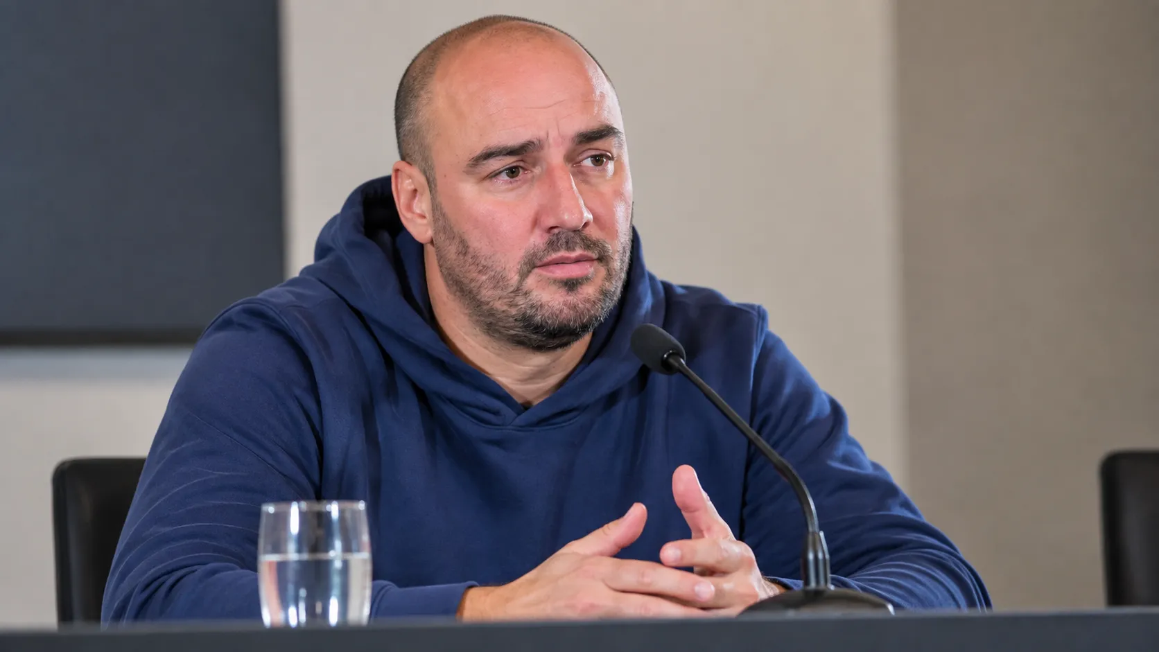 Stijn Stijnen spreekt in een blauwe hoodie aan een tafel tijdens een persconferentie.