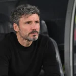 Een geconcentreerde coach in een zwarte trui zit in de dug-out.