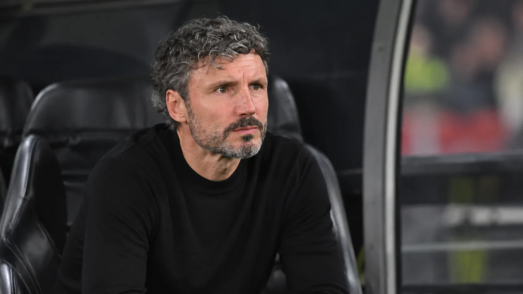 Een geconcentreerde coach in een zwarte trui zit in de dug-out.