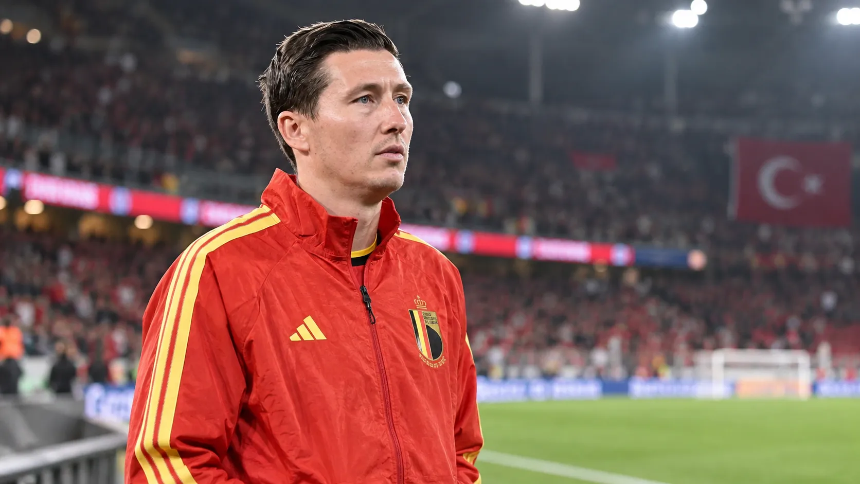 Hans Vanaken in een rode trainingsjas van België aan de zijlijn van een verlicht stadion.