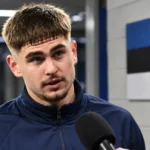 Maksim Paskotši in een neutrale trainingsjas in de mixed zone naast een Ests vlagpaneel.