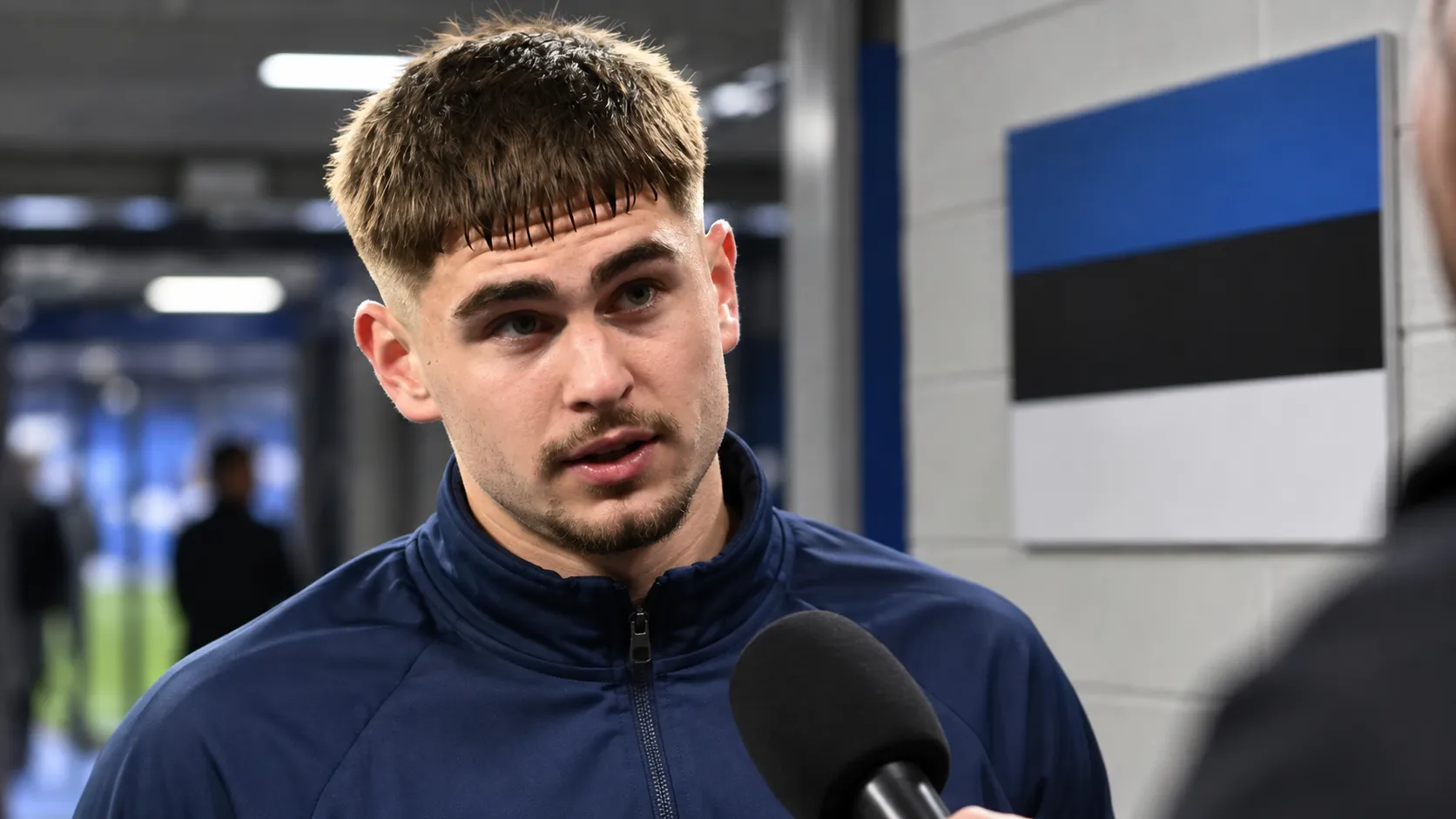 Maksim Paskotši in een neutrale trainingsjas in de mixed zone naast een Ests vlagpaneel.