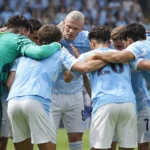 Een groep voetballers in lichtblauwe tenues vormt vlak voor een wedstrijd een kring.
