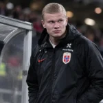 Erling Haaland staat na zijn wissel langs de zijlijn in een Noorse jas.