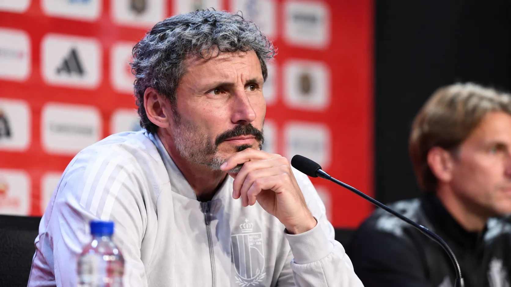Mark van Bommel luistert aandachtig aan een microfoon tijdens een persmoment.