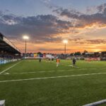 Het Stade de la Cité de l'Oie van URSL Visé met kunstgrasveld, compacte tribunes en oranje-blauwe clubaccenten.