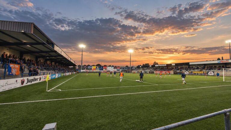 Het Stade de la Cité de l'Oie van URSL Visé met kunstgrasveld, compacte tribunes en oranje-blauwe clubaccenten.