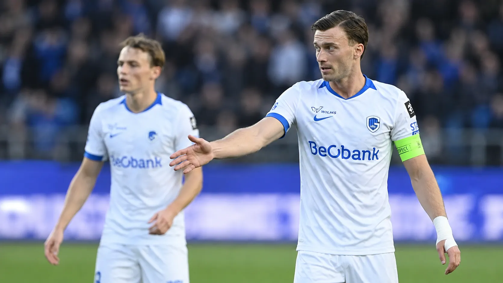 Twee KRC Genk-spelers in witte shirts met blauwe details op het veld.