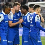 Een groep KAA Gent-spelers in blauw-witte wedstrijdtenues verzamelt zich op het veld.