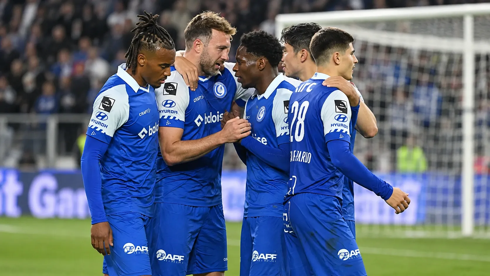 Een groep KAA Gent-spelers in blauw-witte wedstrijdtenues verzamelt zich op het veld.