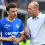 Een speler in blauw tenue en een coach overleggen naast het veld.