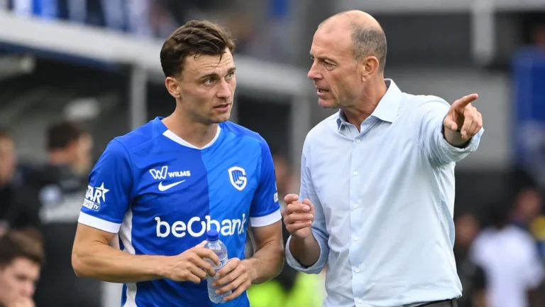 Een speler in blauw tenue en een coach overleggen naast het veld.