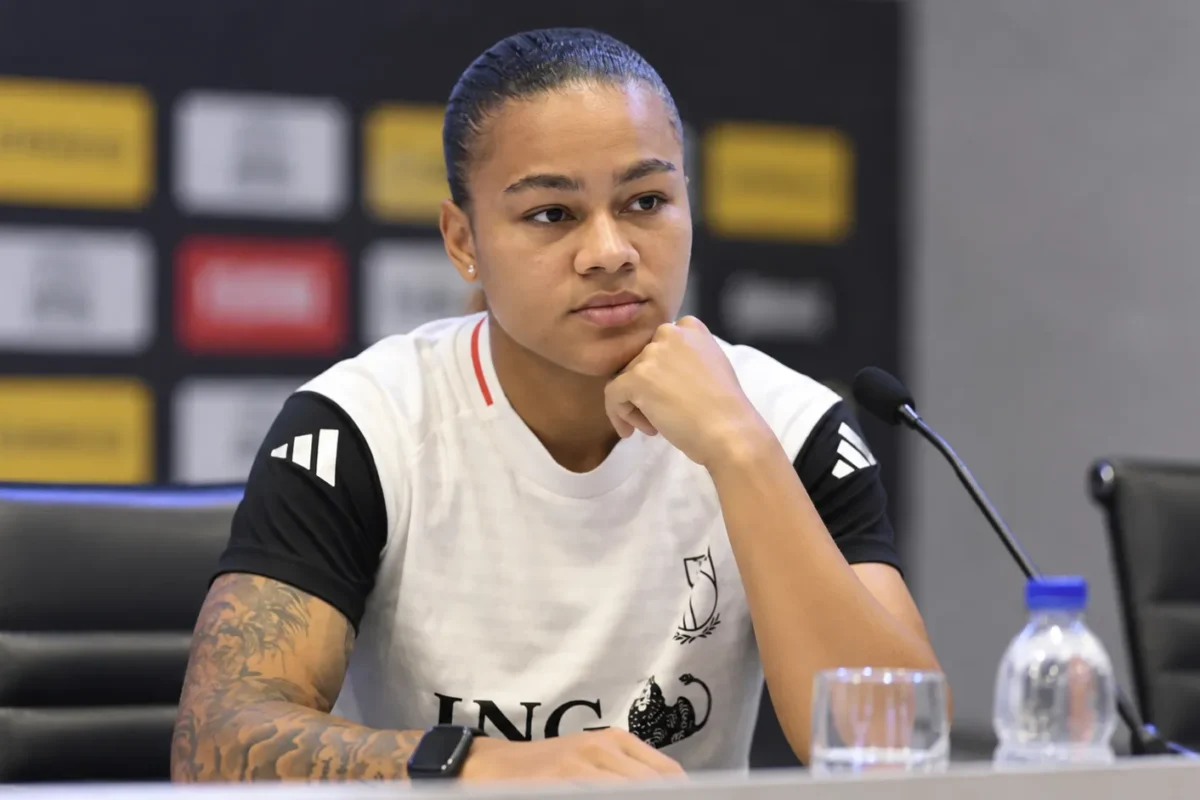 Een vrouwelijke voetbalster zit aan een persconferentietafel in een wit-zwart sportshirt.
