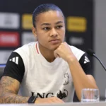 Een vrouwelijke voetbalster zit aan een persconferentietafel in een wit-zwart sportshirt.