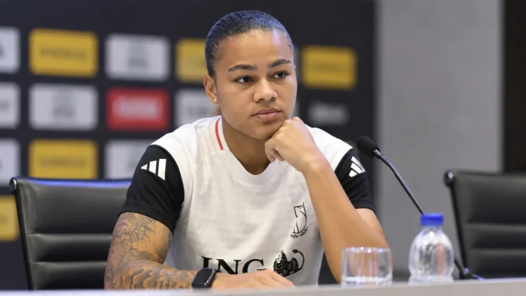 Een vrouwelijke voetbalster zit aan een persconferentietafel in een wit-zwart sportshirt.
