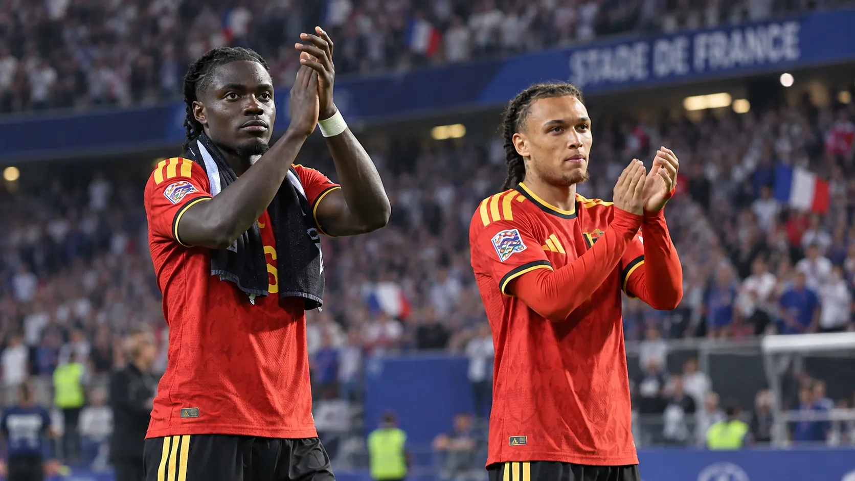 Twee Belgische internationals in rood tenue applaudisseren langs het veld.