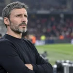 Een niet-geïdentificeerde coach staat geconcentreerd langs de zijlijn van een voetbalveld.