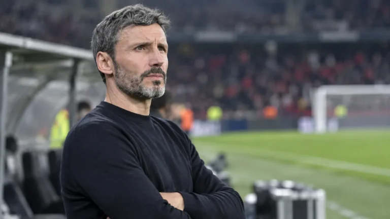 Een niet-geïdentificeerde coach staat geconcentreerd langs de zijlijn van een voetbalveld.