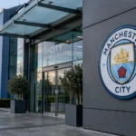 De ingang van een Manchester City-clubgebouw met het clubembleem op een vaste gevelplaat.