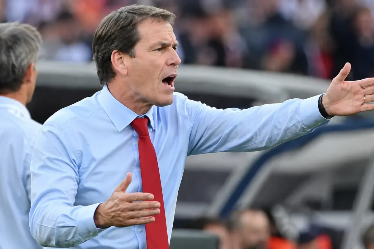 Een niet-spelende coach in hemd en das gebaart langs de zijlijn van een voetbalstadion.