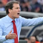 Een niet-spelende coach in hemd en das gebaart langs de zijlijn van een voetbalstadion.