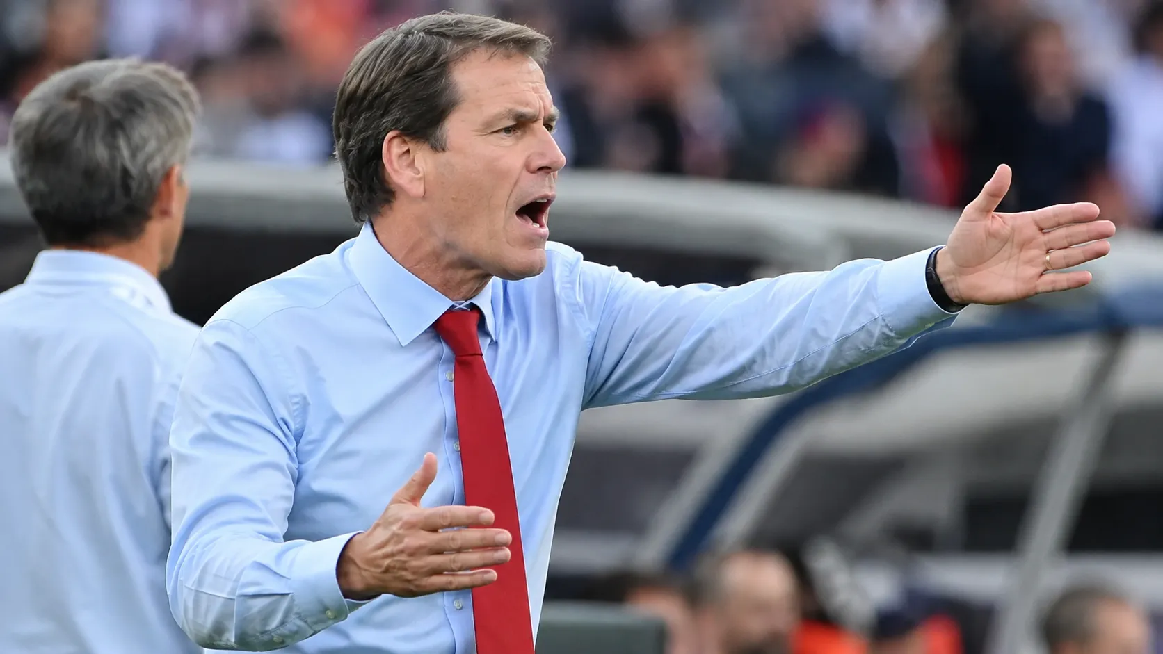Een niet-spelende coach in hemd en das gebaart langs de zijlijn van een voetbalstadion.