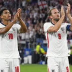 Twee Engelse voetballers in witte shirts applaudisseren richting de tribunes na een wedstrijd.
