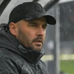 Stijn Stijnen in zwarte pet en regenjas aan de rand van een voetbalveld.