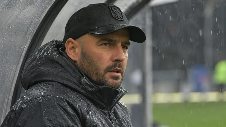 Stijn Stijnen in zwarte pet en regenjas aan de rand van een voetbalveld.