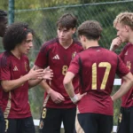 Een groep jonge voetballers in donkerrode Belgische tenues staat samen langs het veld.