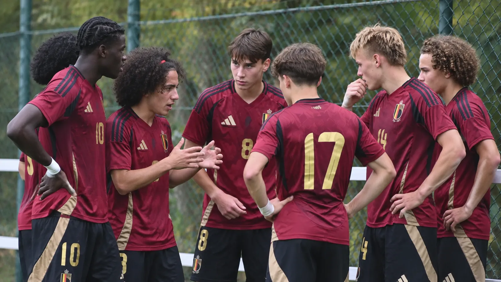 Een groep jonge voetballers in donkerrode Belgische tenues staat samen langs het veld.