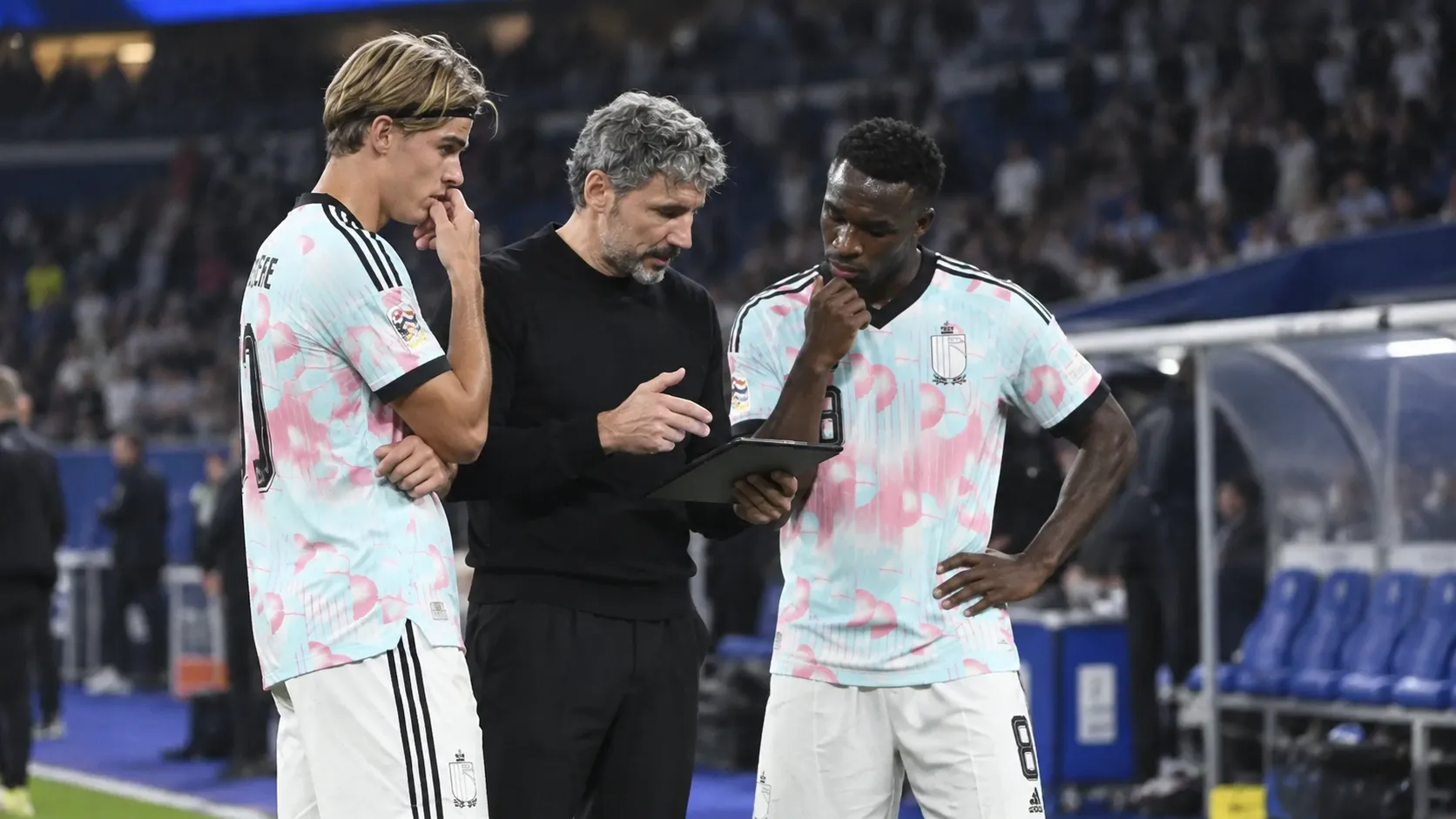 Een staflid spreekt langs de zijlijn met twee voetballers in lichte shirts.