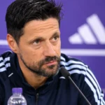Een trainer in een donker trainingsjack zit aan tafel in een paarse persruimte.