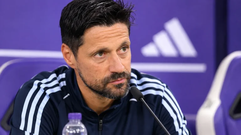Een trainer in een donker trainingsjack zit aan tafel in een paarse persruimte.
