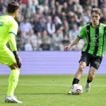 Een speler in een groen-zwart tenue in duel met een doelman in een neon geel tenue.