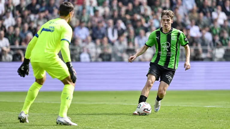 Een speler in een groen-zwart tenue in duel met een doelman in een neon geel tenue.