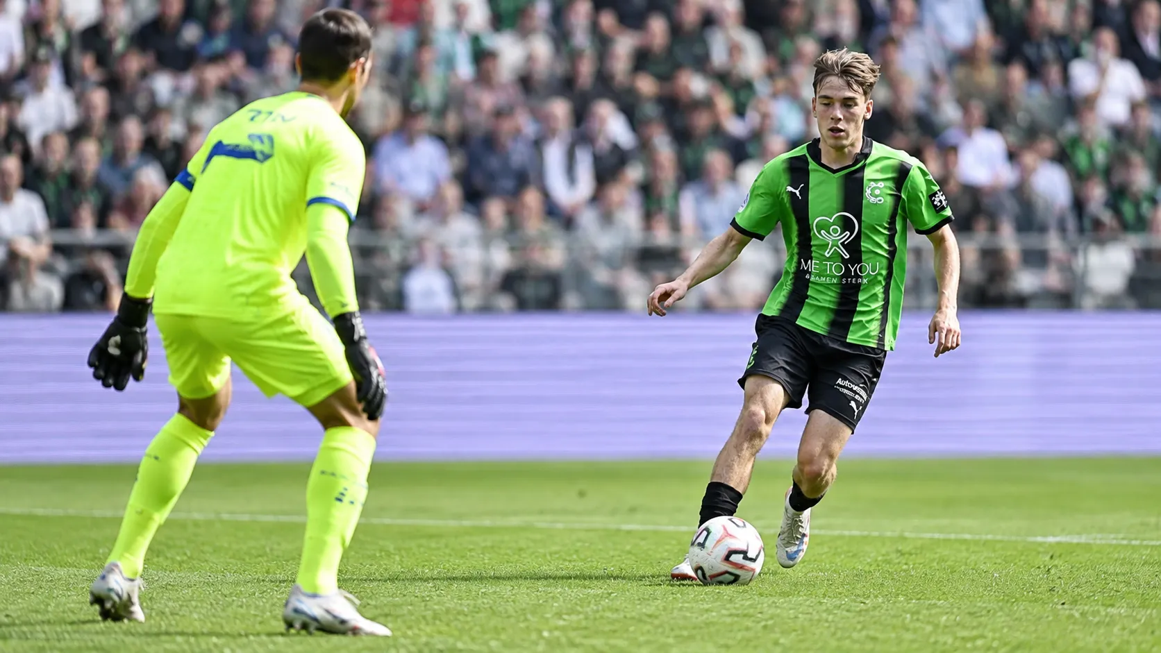 Een speler in een groen-zwart tenue in duel met een doelman in een neon geel tenue.