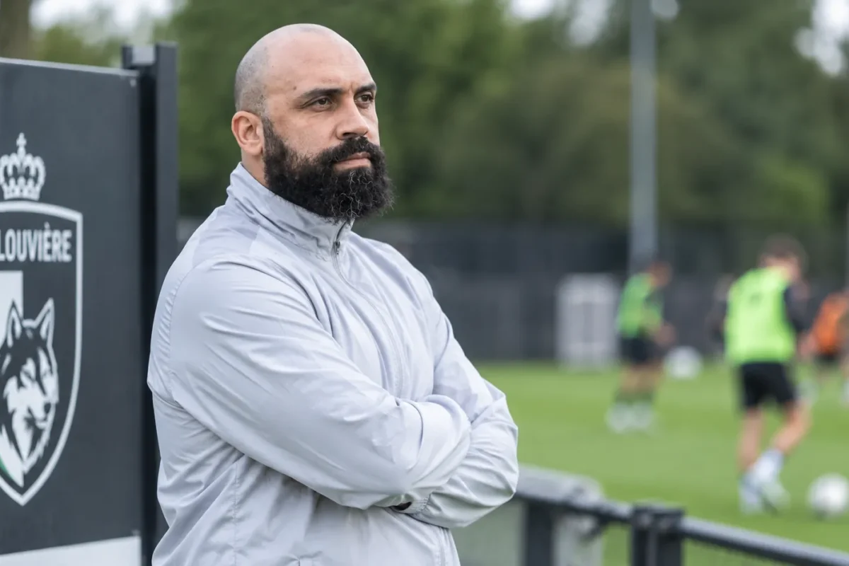 Anthony Vanden Borre kijkt langs een trainingsveld van La Louvière, met een clubembleem op een vast bord naast het veld.