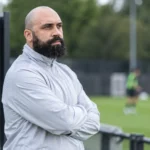 Anthony Vanden Borre kijkt langs een trainingsveld van La Louvière, met een clubembleem op een vast bord naast het veld.